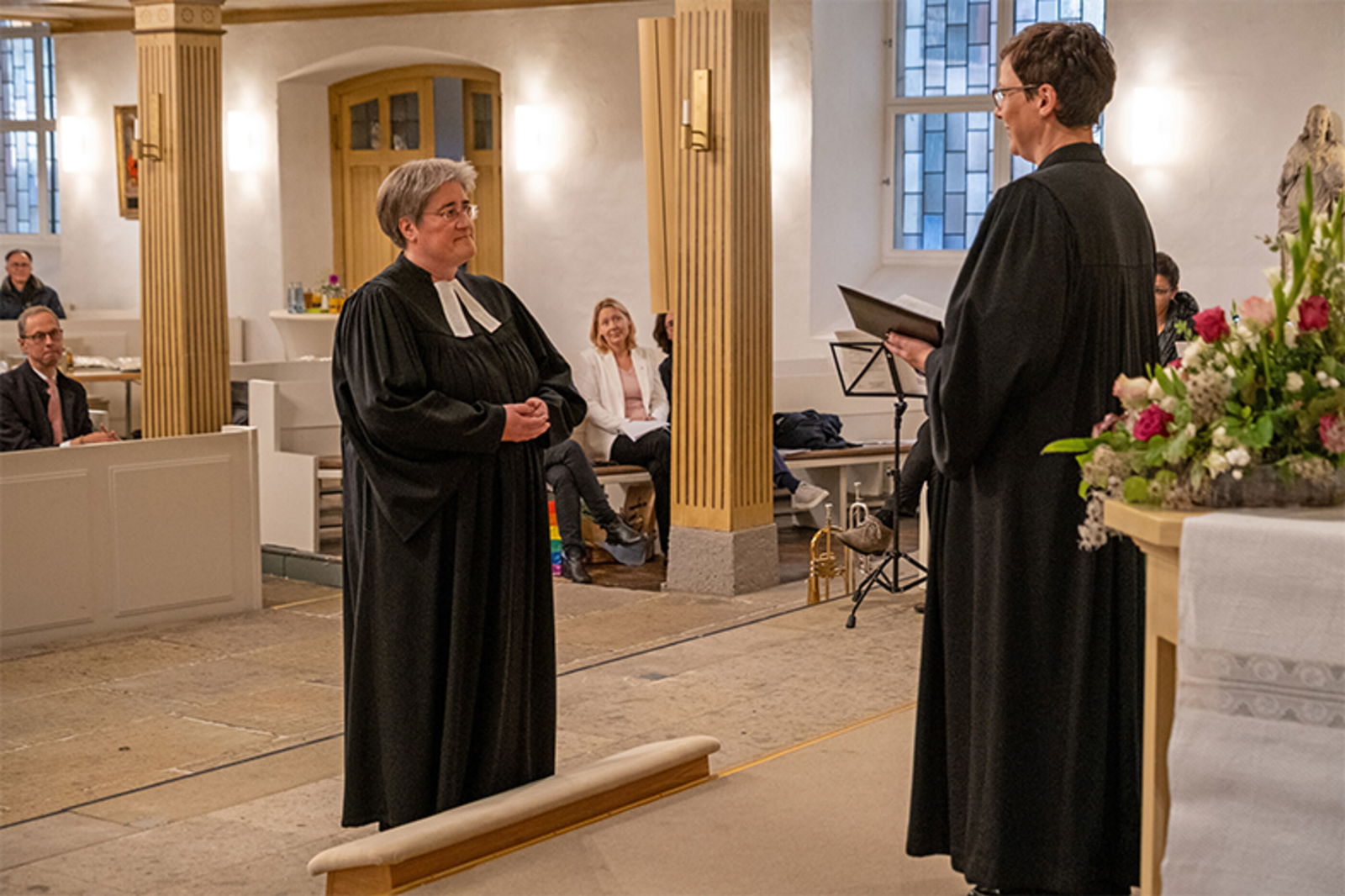Zwei weiblich gelesene Personen in Talaren stehen in einer Kirche voreinander.