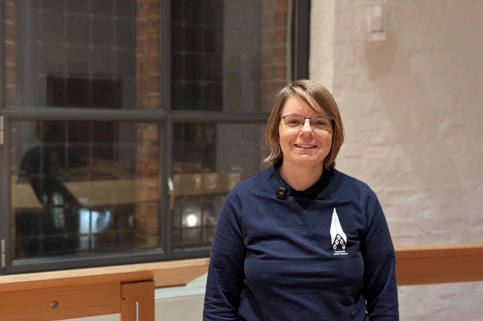 Eine weiblich gelesene Person in blauem Pulli vor einem Kirchenfenster.