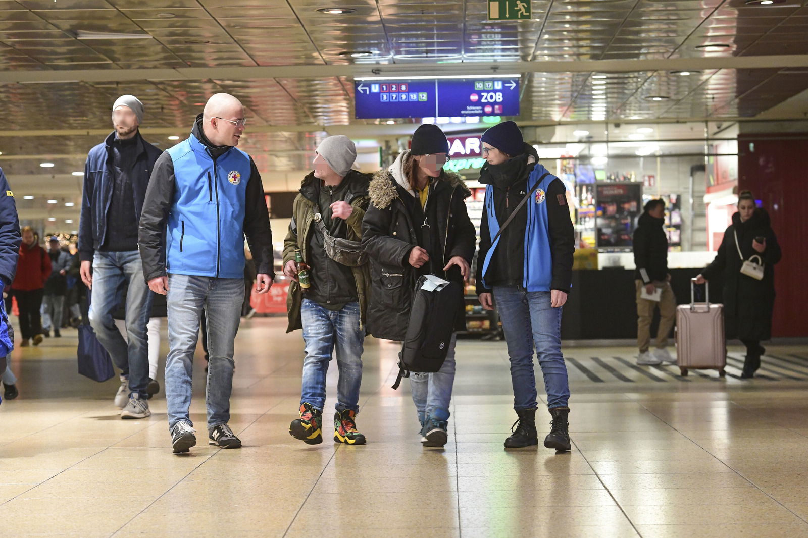 Zwei Personen in blauen Westen begleiten zwei Menschen im Bahnhof.