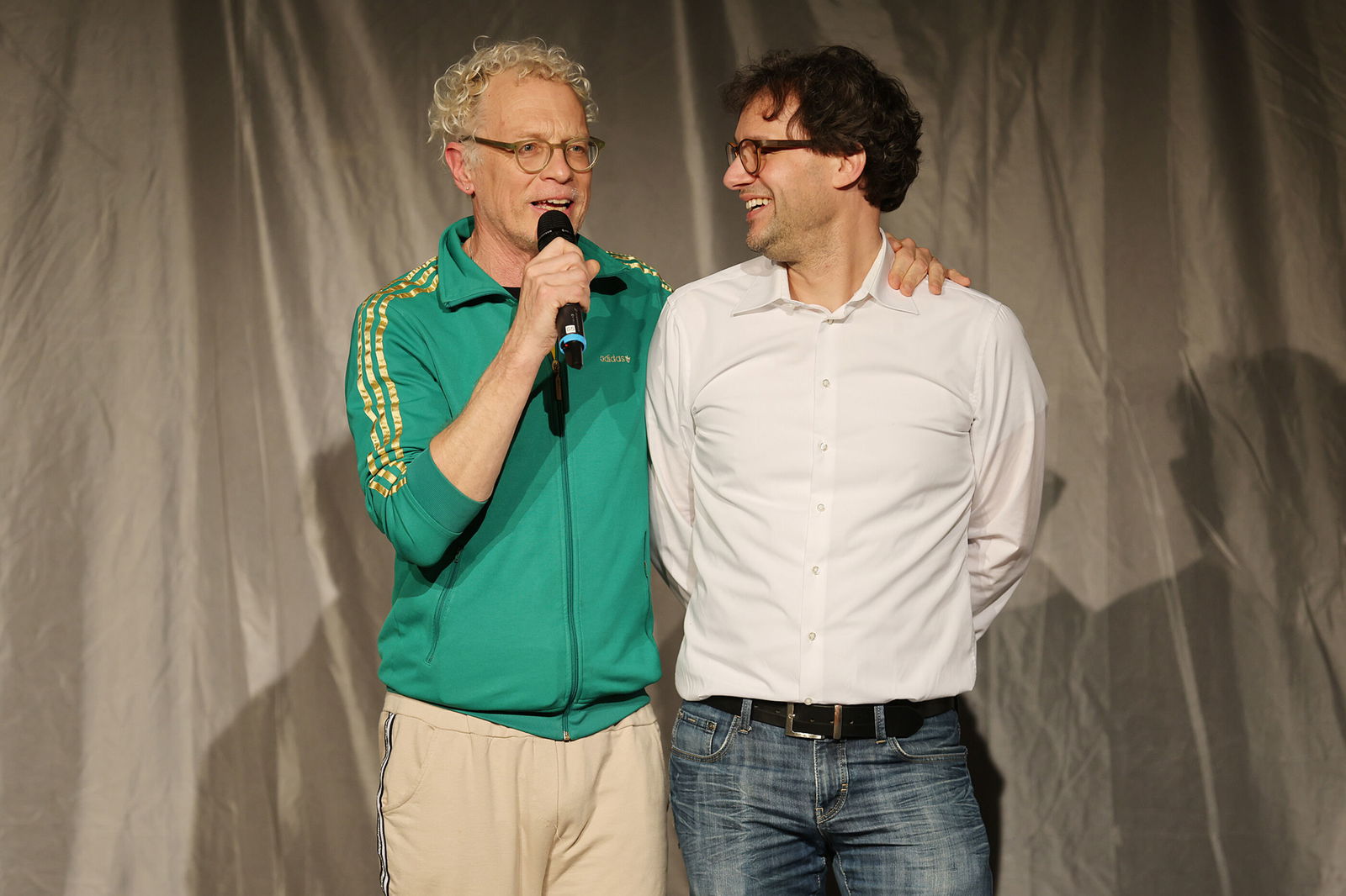 Zwei als Männer lesbare Personen unterhalten sich mit einem Mikrophon auf einer Bühne.