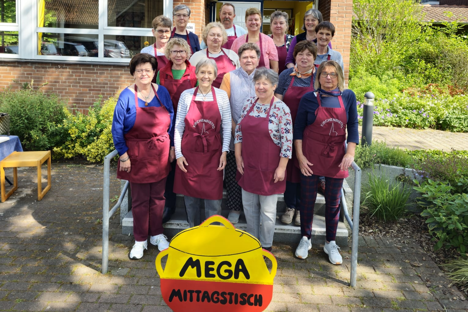 Ein dutzend Personen, weiblich gelesen, stehen zusammen vor einem Haus und hinter einem gemalten Suppentopf, auf dem "Mega Mittagstisch" steht.