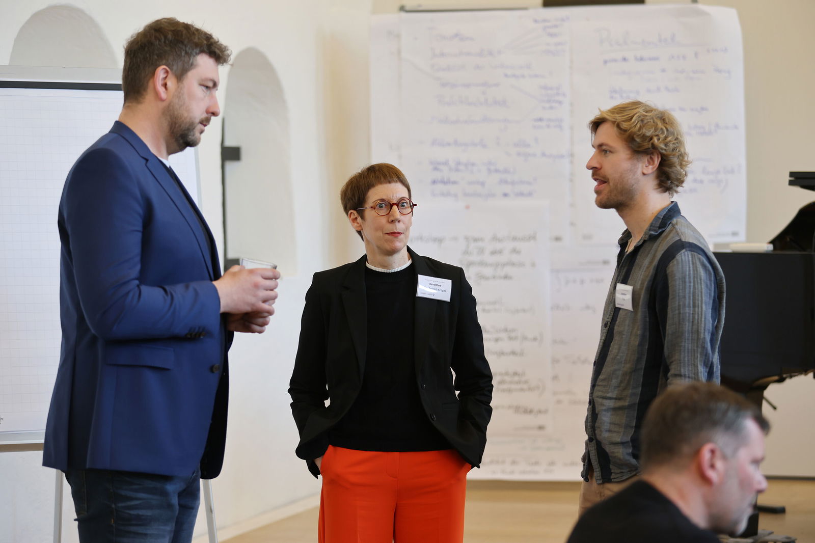 Zwei als Männer und eine als Frau lesbare Personen unterhalten sich bei einer Tagung.