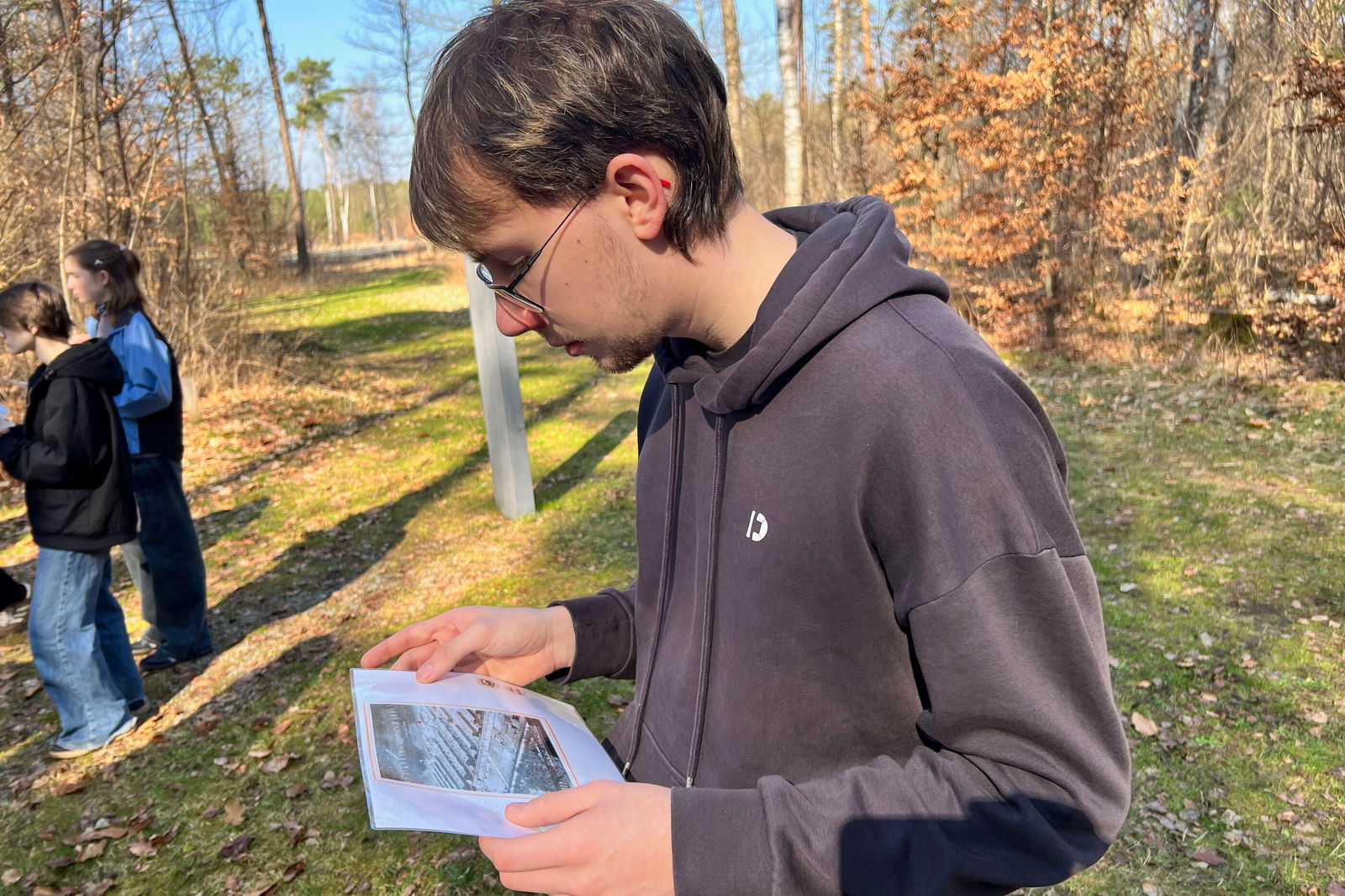 Eine junge, männlich gelesene Person schaut ein schwarz-weißes Bild an. Sie steht im Freien.