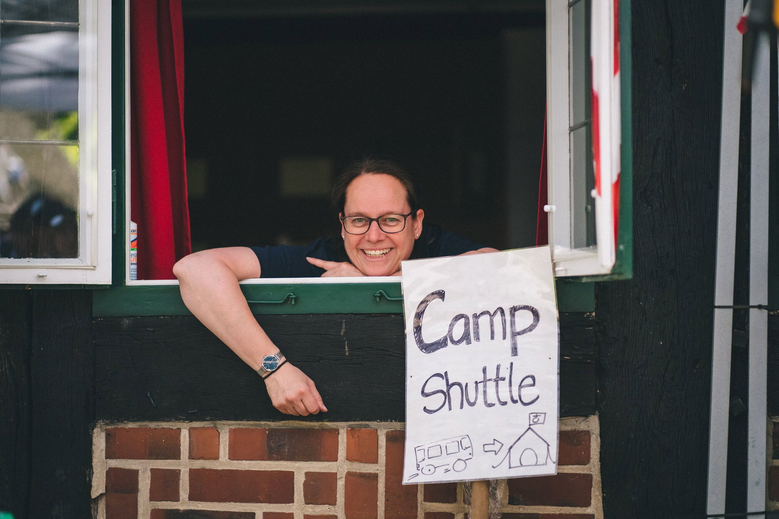 Eine als Frau lesbare Person schaut aus einem Fenster, unter ihr ein handgeschriebenes Blatt mit der Aufschrift: Camp-Shuttle.