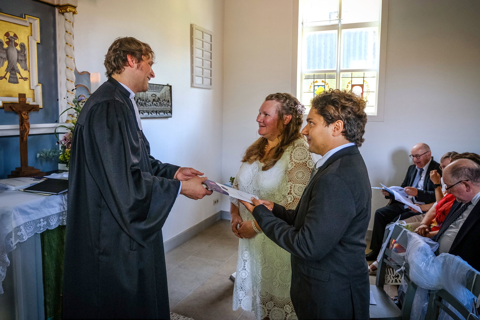 Ein Pastor verheiratet vor einem Altar in einer Kirche eine als Mann und eine als Frau lesbare Person.