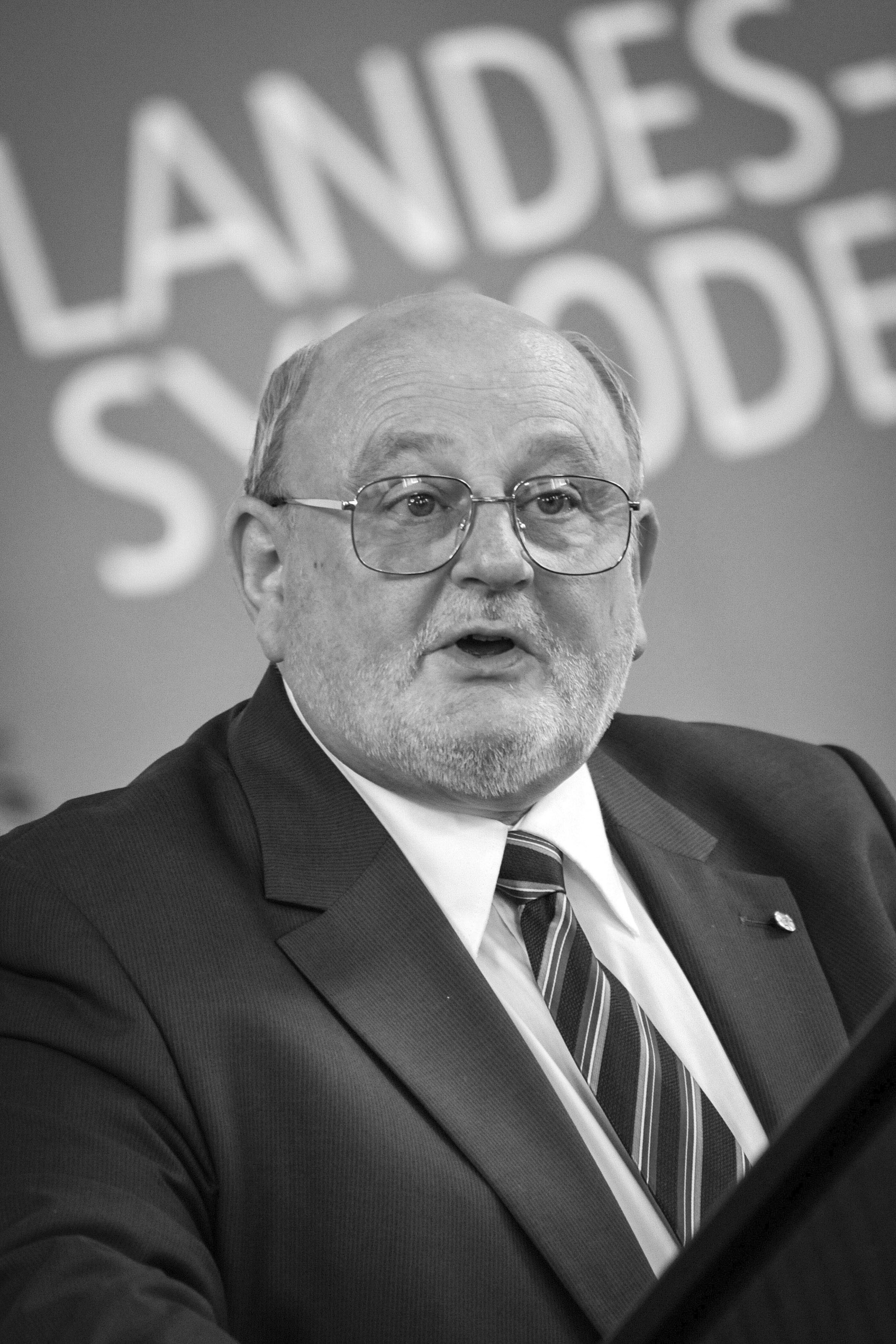 Foto einer als Mann lesbaren Person mit Brille und hoher Stirn hinter einem Mikrofon. Im Hintergrund steht Landessynode auf einem Banner.