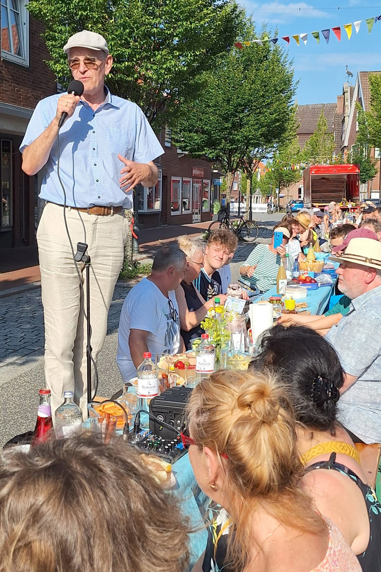 Eine als Mann lesbare Person mit Schiebermütze steht mit Mikrofon auf einer Bank und hält an einer langen Tafel im Freien eine Rede.