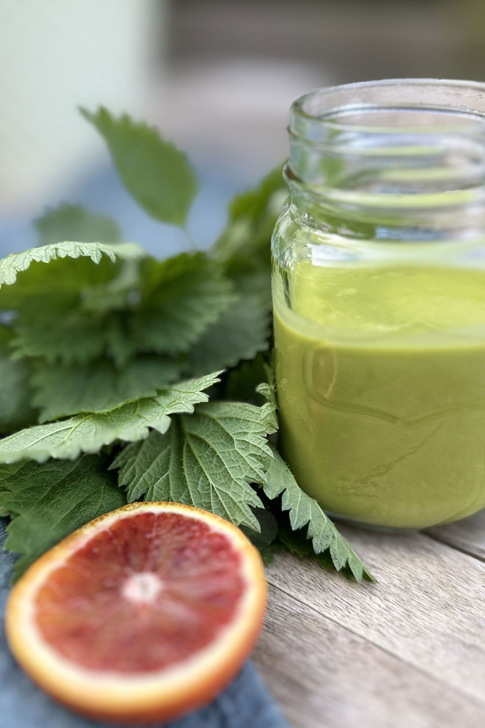 In einem Glas steht grüne, dickliche Flüssigkeit, ein Smoothie. Daneben liegen Brennesseln und eine halbe Blutorange auf einem Tisch.