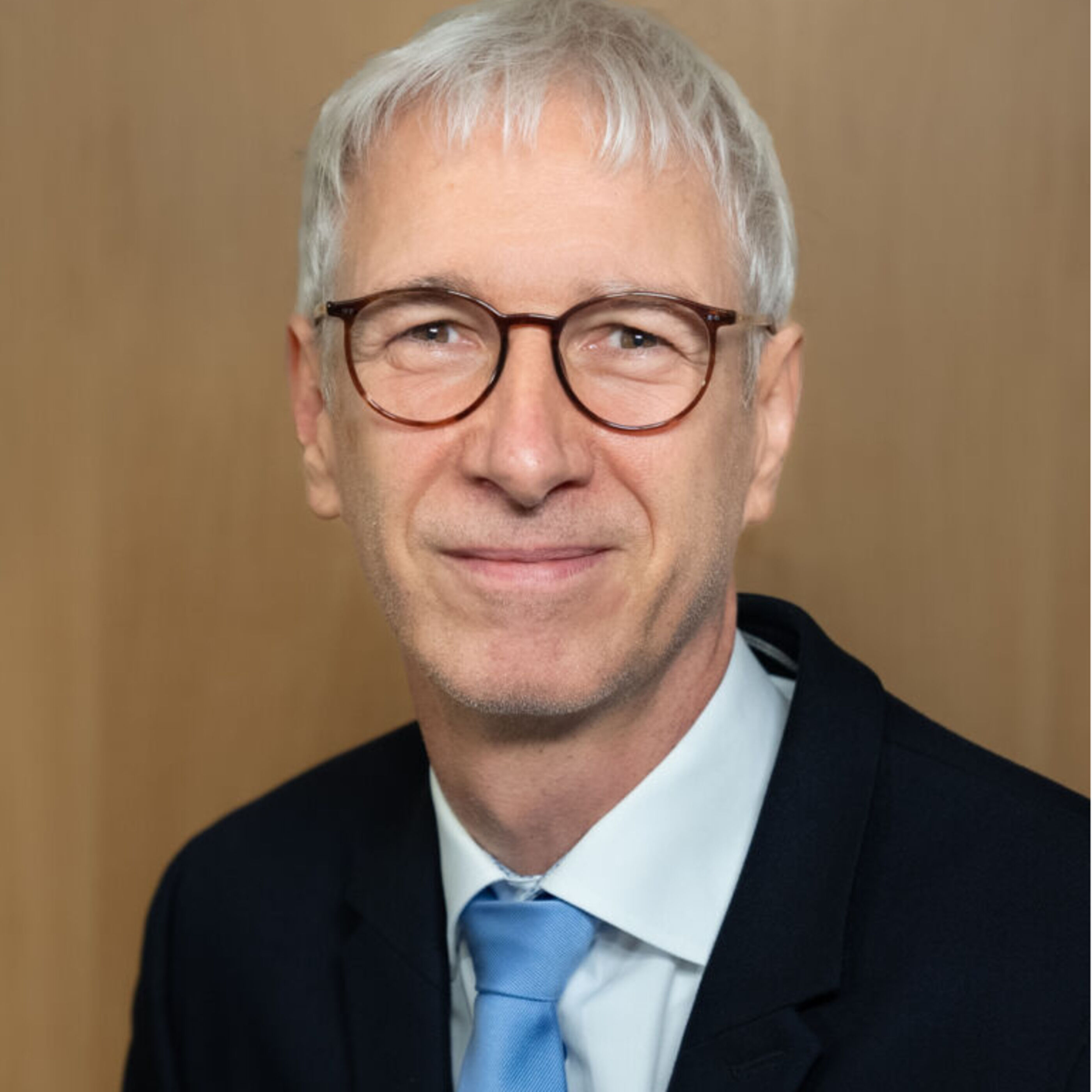 Ein Man lächelt in die Kamera. Er trägt einen schwarzen Anzug und eine blaue Krawatte. Der Mann hat graue, kurze Haare und trägt eine braune Hornbrille.