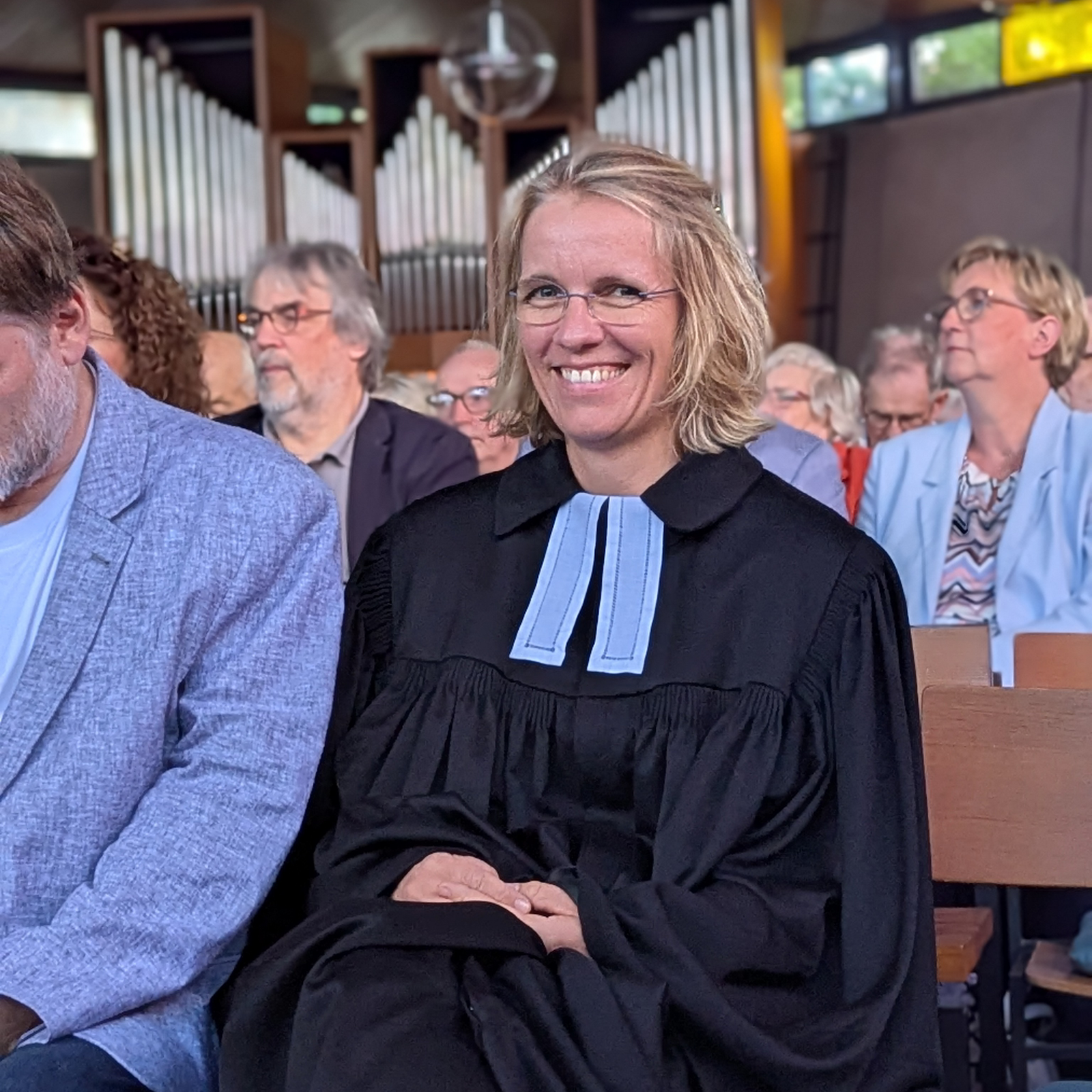 Eine weiblich gelesene Person im Talar sitzt breit lächelnd in einer Kirche.
