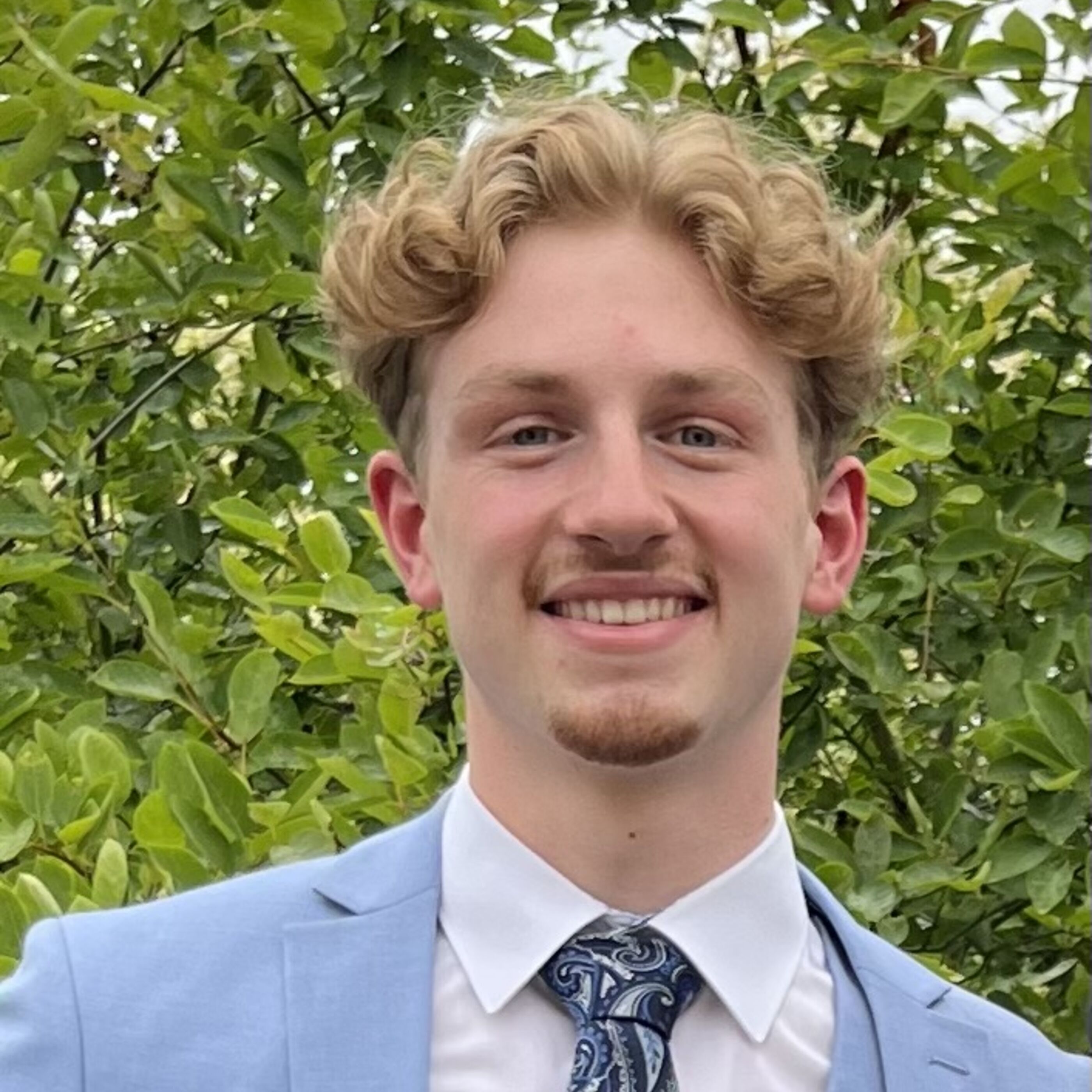 Ein junger Mann mit blonden Locken und Bart trägt einen blauen Anzug.