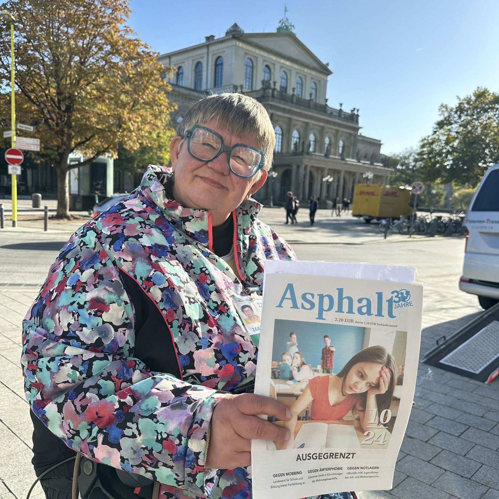 Eine weiblich gelesene Person mit bunter Jacke und Brille sitzt auf einem Rollator und hält ein Straßenmagazin in Händen. Im Hintergrund ist die Oper Hannover erkennbar.