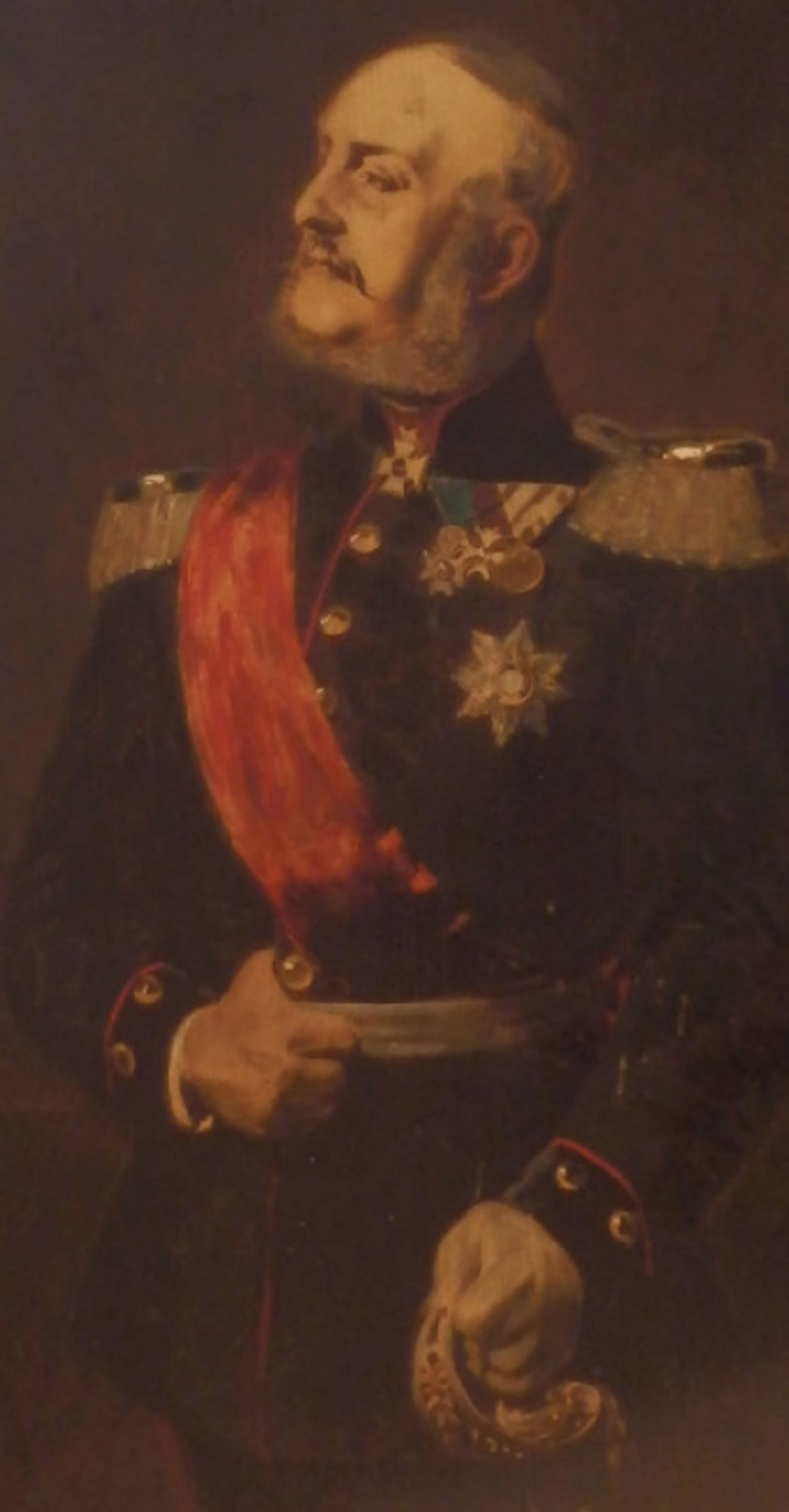 Das Bild zeigt ein Ölgemälde von König Georg V., eine männlich lesbare Person in Uniform mit kurzen grauen Haaren, einem Schnurbart, mit Schärpe und Orden.