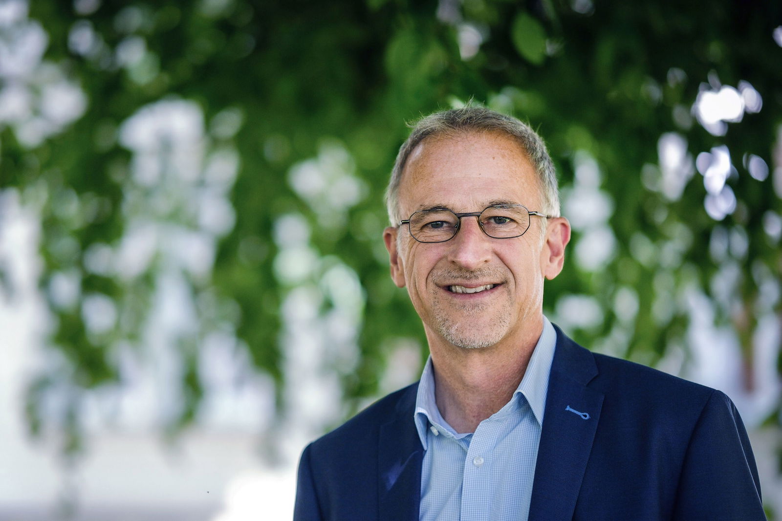 Eine männlich gelesene Person mit kurzen grauen Haaren, Brille und kurzem Bart steht unter einem Baum. Er trägt ein dunkelblaues Sakko und ein hellblaues Hemd. 