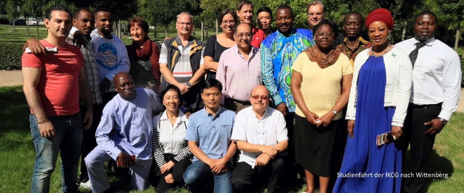 Eine Gruppe von Menschen unterschiedlicher Hautfarbe hat sich draußen auf einer Rasenfläche zum Gruppenfoto aufgestellt.
