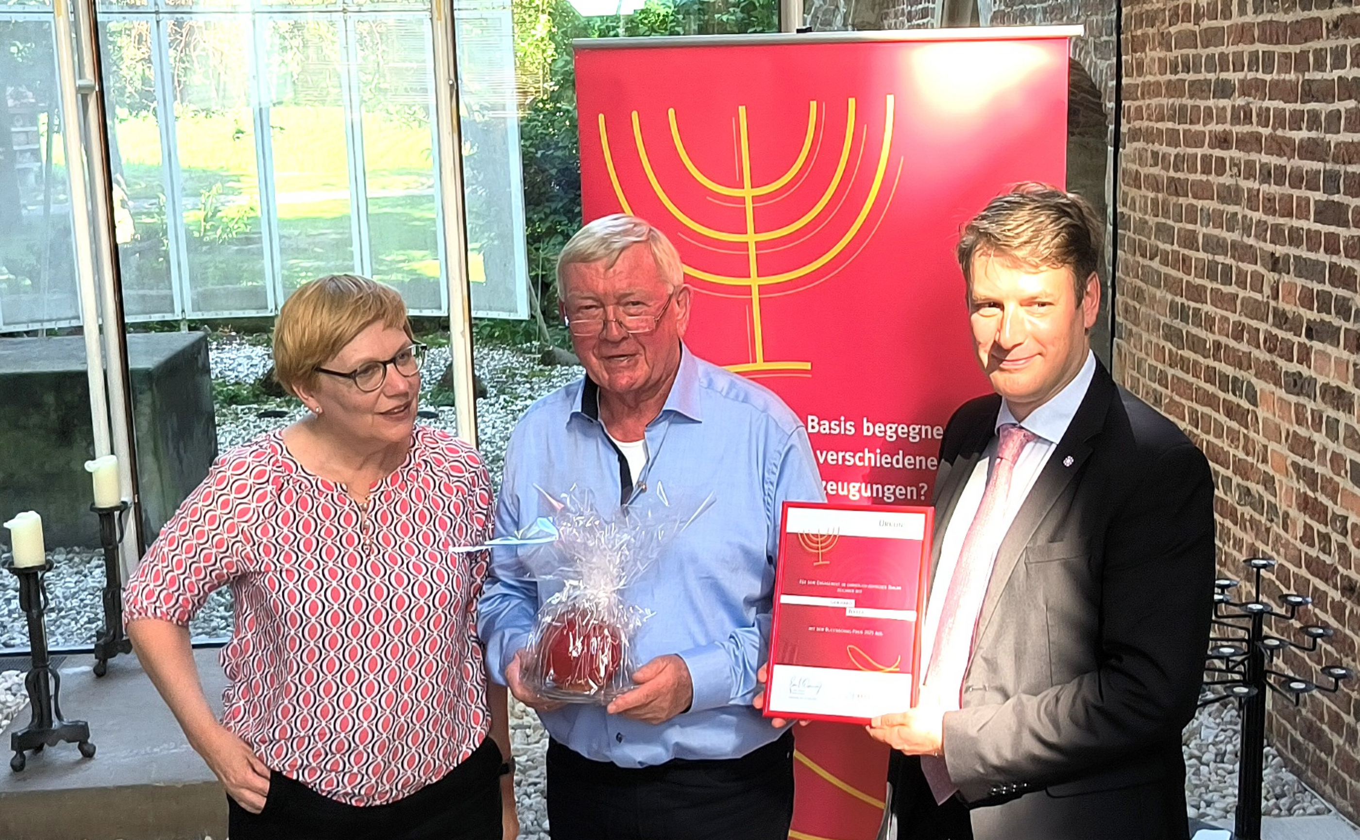 Eine weiblich und zwei männlich gelesene Personen stehen zusammen. Einer hält ein Geschenk, ein anderer eine Auszeichnung.