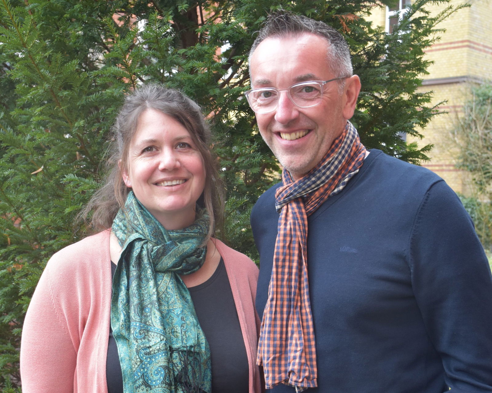 Eine weibliche und eine männlich gelesene Person mit Schals stehen nebeneinander.