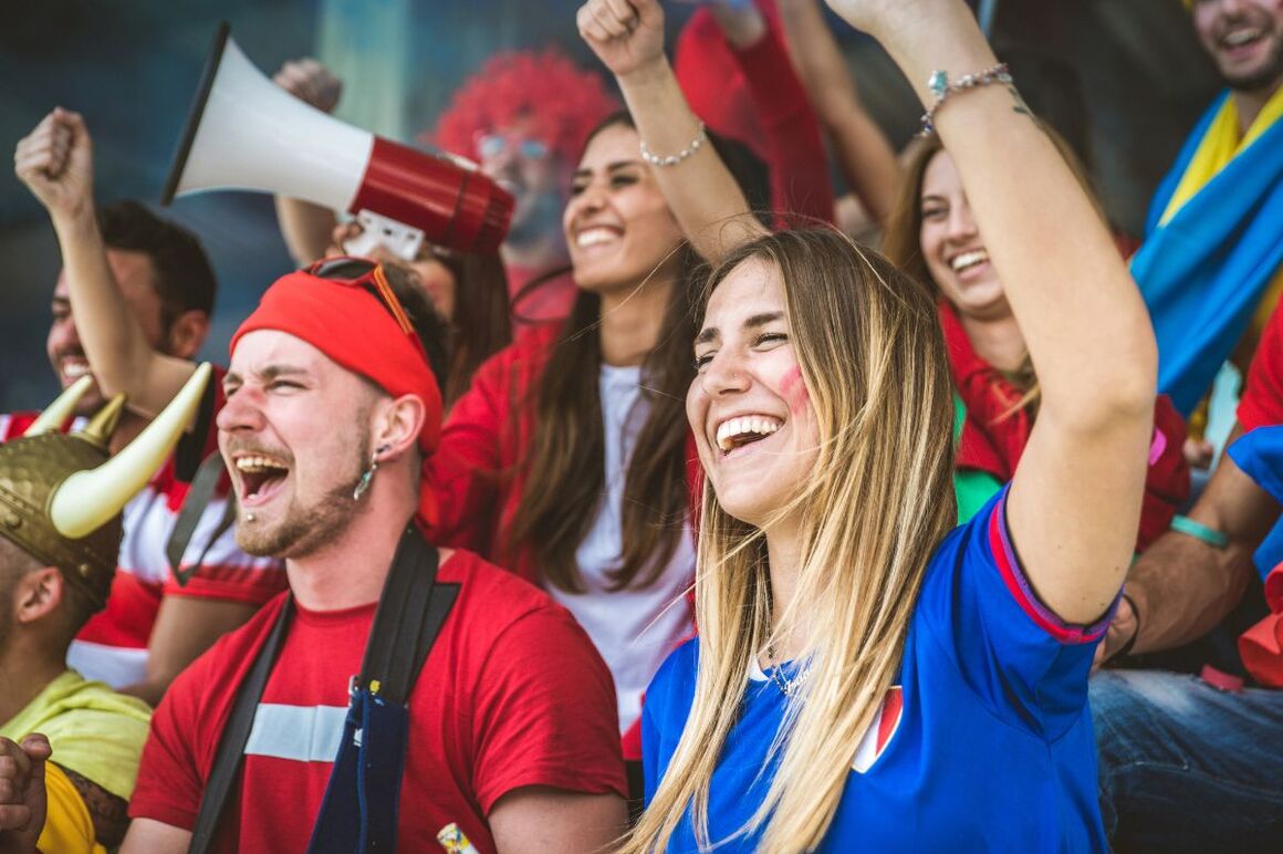 Weiblich und männliche gelesene Personen mit bunten Trikots und Accessoires stehen auf einer Tribüne und jubeln.