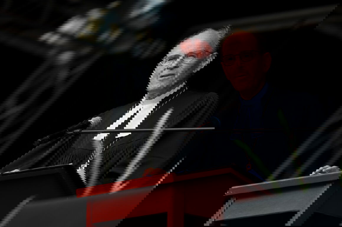 Eine mittelalte als Mann lesbare Person steht im Talar an einem Redepult und spricht.