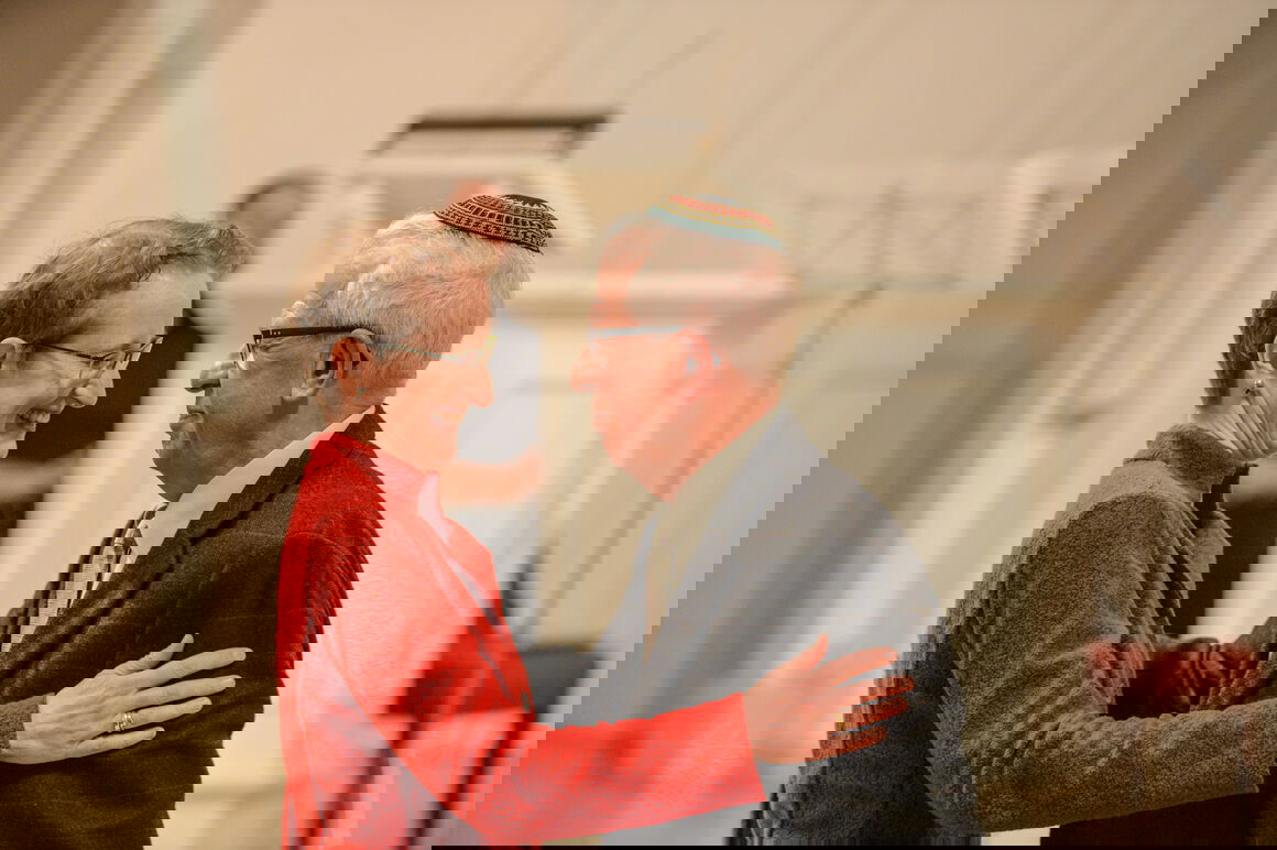 Eine Frau mit kurzen Haaren und roter Jacke hält einen Mann mit Kippa an den Armen.