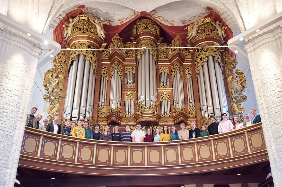 Blick hinauf zu einer Orgel auf einer Empore. An der Balustrade stehen etwa zwei Dutzend junge Menschen und schauen herunter.