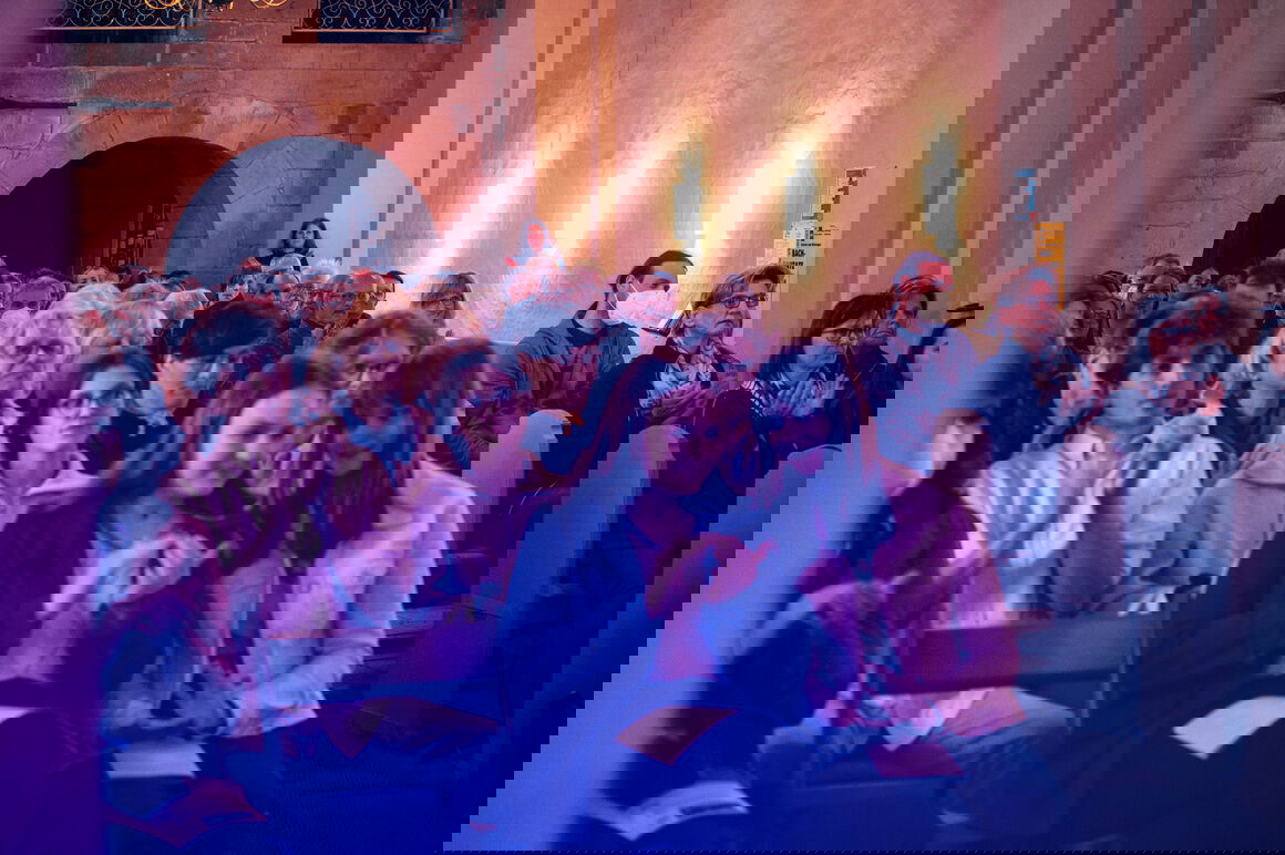 Viele, vor allem junge Menschen sitzen in einem Raum mit hohen Wänden, evt. einer Kirche.