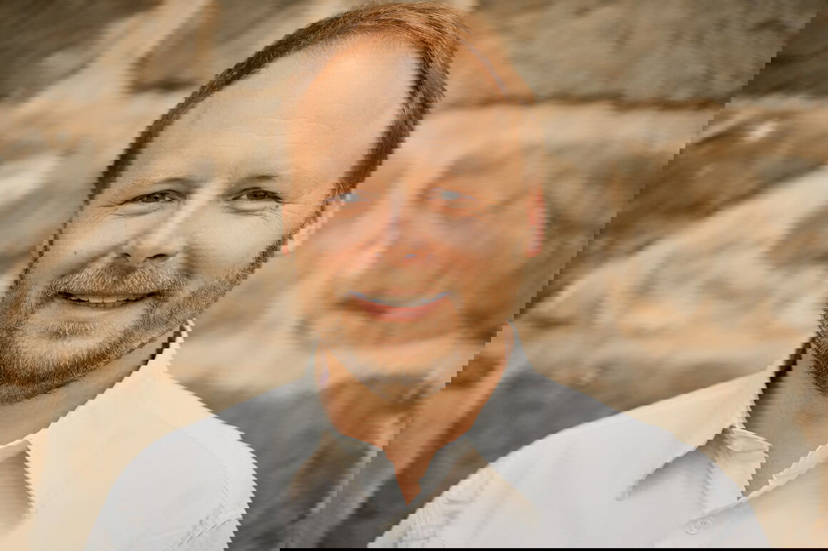Eine männlich gelesene Person in weißem Hemd lächelt freundlich vor einer Steinwand.