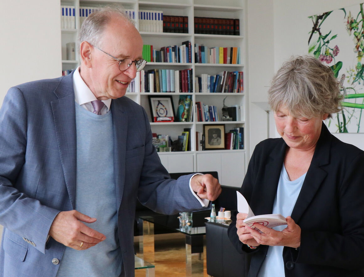 Eine als Mann und eine als Frau lesbare Person stehen in einem Büro uns sprechen über ein kleines Büchlein.