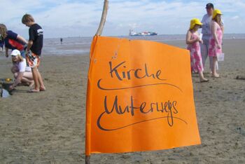 Eine orangefarbene Flagge steht auf einem Strand. Darauf steht: Kirche unterwegs. Im Hintergrund sieht man Menschen und das Meer.