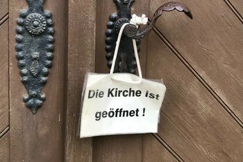 An einer Kirchentür hängt ein Schild. Darauf steht: Die Kirche ist geöffnet
