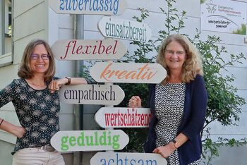 Auf dem Bild sind zwei Mitarbeitende des Dorfhelferinnenseminars zu sehen. Zwischen ihnen stehen Schilder mit Wörtern wie achtsam, flexibel, geduldig, empathisch und weitere.