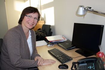 Eine Frau sitzt am Schreibtisch vor einem Computer. Sie trägt eine graue Strickjacke und eine Brille und hat braune, kurze Haare. Sie lächelt in die Kamera.