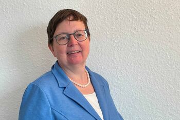 Das Bild zeigt Andrea Schridde. Sie trägt einen hellblauen Blazer und ein weisses Schirt. Sie hat dunkelblonde kurze Haare, trägt eine Brille und eine helle Perlenkette.