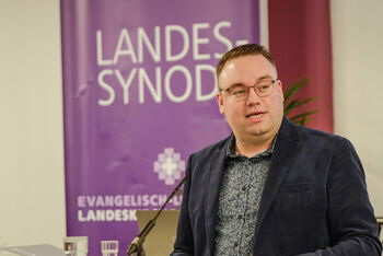 Eine männlich lesbare Person steht an einem Rednerpult vor einem Plenum