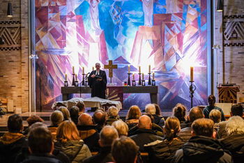 Männlich und weiblich zu lesende Personen sitzen in einem Kirchenraum und schauen auf eine männlich zu lesende Person im Talar, die im Altarraum steht und etwas vorliest.