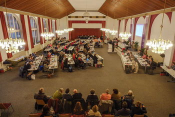 Weiblich und männlich zu lesende Personen sitzen im Tagungsraum der Landessynode an Tischen. Am Pult steht eine männlich zu lesende Person.