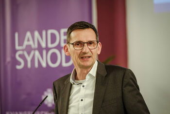 Eine männlich gelesene Person mit dunklen, kurzen Haaren und Brille. 