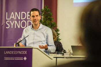 Eine männlich lesbare Person im Hemd steht an einem Pult und spricht.