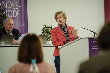 Eine weiblich lesbare Person mit Blazer, Rollkragenpullover und Brille steht an einem Pult und spricht.