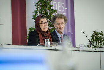 Eine weiblich lesbare Person mit islamischem Kopftuch, eine weiblich lesbare Person mit Schal und eine männlich lesbare Person im Anzug sitzen nebeneinander