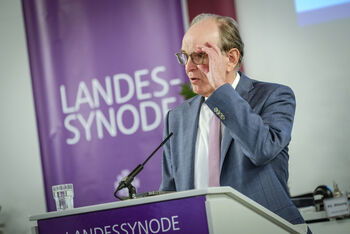 Eine männlich lesbare Person mit Brille und kurzen Haaren steht im Anzug an einem Pult, fasst sich an die Brille  und redet. 