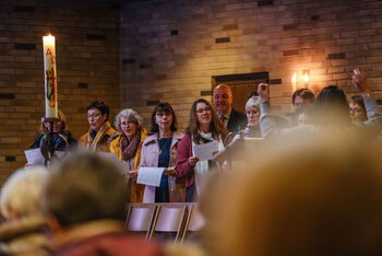 Das Bild zeigt eine singende Gruppe von Personen mit Liedzetteln in den Händen, die neben einer großen Kerze stehen.