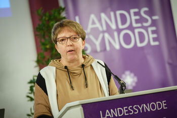 Eine weiblich lesbare Person mit Brille und kurzen Haaren steht hinter einem Rednerpult und spricht.