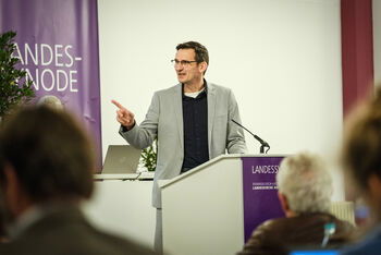 Eine männlich lesbare Person mit Brille und dunklen Haaren steht im Anzug hinter einem Rednerpult und spricht in ein Mikrofon.