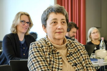 Eine weiblich lesbare Person mit karierter Bluse sitzt, mehrere weiblich lesbare Personen im Hintergrund