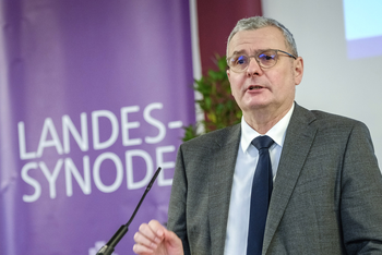 Eine männlich lesbare Person steht an einem Rednerpult mit Mikrofon