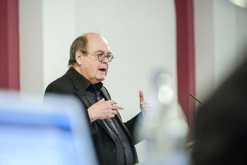 Eine männlich lesbare Person  mit Brille steht und gestikuliert 