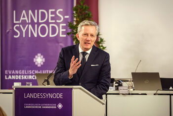 Eine männlich lesbare Person steht an einem Rednerpult und hebt eine Hand und spricht lächelnd zu den Zuhörerinnen.