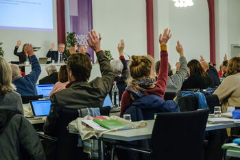 Das Bild zeigt eine Gruppe von Menschen von hinten, die an Tischen sitzen und für eine Abstimmung die Hände heben.