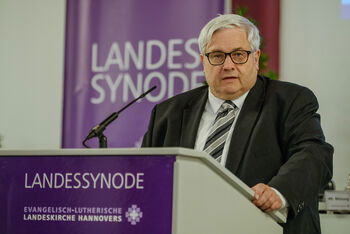 Eine männlich lesbare Person mit einer Brille steht an einem Rednerpult und spricht.